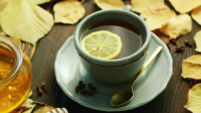 From above view of cup of hot tea with slice of lemon and bowl of honey placed near decorated with dry leaves on wooden background