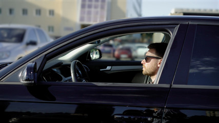 A young man with a beard rides in the parking lot on his black car. Business sedan quietly moves along other machines.