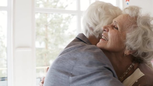 Happy elderly woman embracing with female guest and saying thanks while friends congratulating her at birthday party