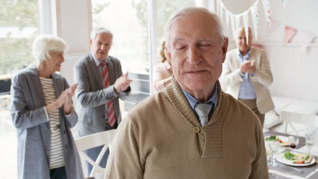 Cheerful senior man standing by table with holiday dinner and smiling at camera while his best friends clapping hands at birthday party in living room