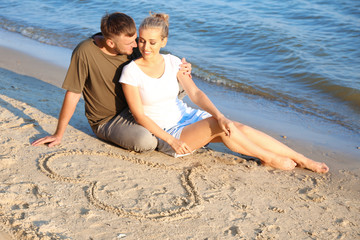Fototapeta na wymiar Cute young couple sitting near heart drawn on beach sand