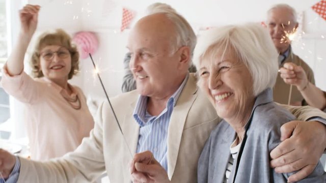 Smiling elderly man and woman embracing and dancing with burning sparklers at party with friends