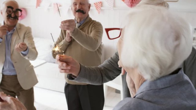 Cheerful senior men and women holding party masks on sticks and sparklers while dancing together at celebration in the living room