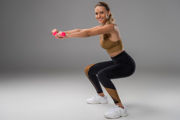 athletic smiling young woman doing squats with dumbbells on grey