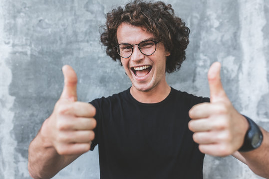 Horizontal shot of handsome happy smiling male with curly hair, wears sepctacles posing for social advertisement with thumbs up on gray concrete wall with copy space for your text.