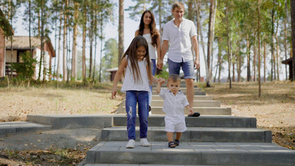 Obraz premium little girl is holding hands of her baby brother, walking together on stone stair in a park in daytime, parents are behind