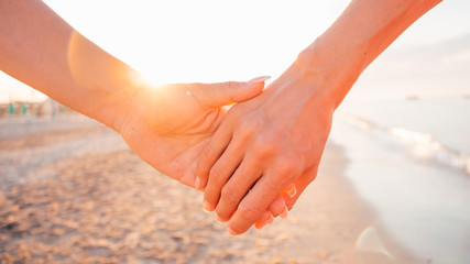 Close up of two female holding hands against sunset - two girls walking on the beach hand by hand -...