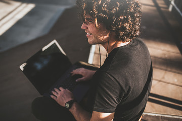 Rear view shot of male with curly hair using laptop for chatting online with friends and earphones to listening the music, connected to free wireless. Handsome man texting messages.