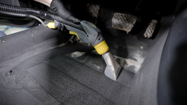washerman is hoovering automobile carpet, cleaning by foam interior of car in a garage, wearing gloves