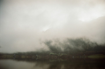 Cottages and villas seen through the dense fog covering water of lake. Shadows of the trees reflected in water. Sleepy misty landscape. Film photography