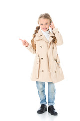 adorable kid in beige coat talking on smartphone and showing something, isolated on white