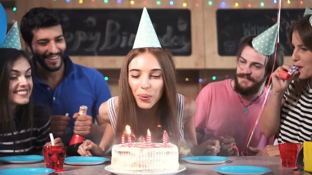 Excited pretty woman blowing birthday candles while dearest friends throwing surprise party, slowmotion in stylish modern flat