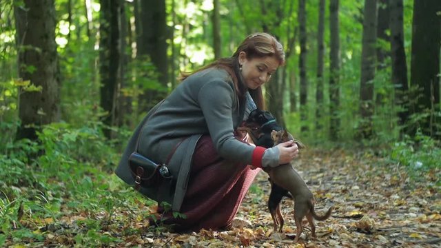 woman in the woods squats and strokes stroking two small dogs.