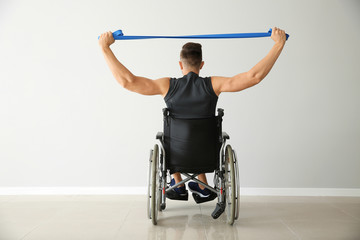 Sporty man training with elastic band while sitting in wheelchair against light wall