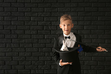 Cute little magician holding hat with rabbit against dark brick wall