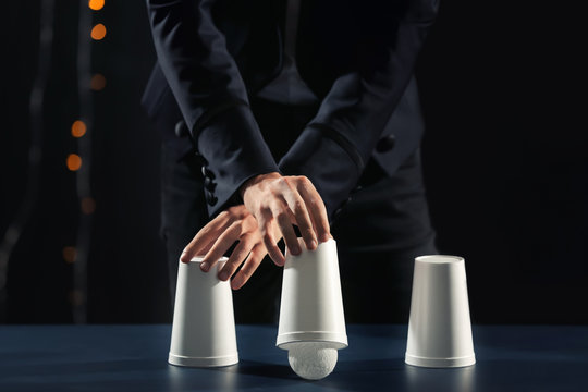 Magician showing tricks with cups on dark background