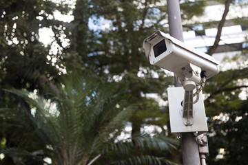 Bellow view of a security camera(cctv) in a public place. With a metal camera, green trees and buildings in the background we compose this photo. Secure, systens, technology and monitoring as concept