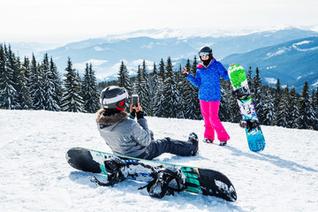 two women in ski suits on top of the mountain do selfie. Girls with snowboards sit on the snow and...