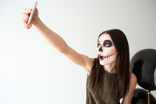 Beautiful woman with Halloween makeup and balloons taking selfie on light background
