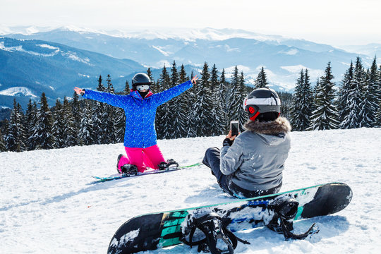 two women in ski suits on top of the mountain do selfie. Girls with snowboards sit on the snow and are photographed