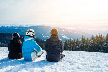Three people in ski suits are sitting on the snow at the top of the mountain and looking into the distance.