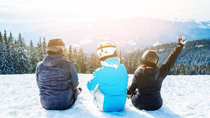 Three people in ski suits are sitting on the snow at the top of the mountain and looking into the...