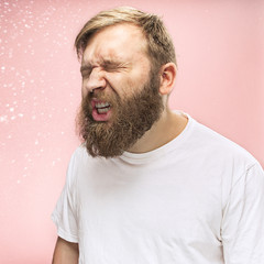 Young funny handsome man with beard and mustache sneezing with spray and small drops, studio...