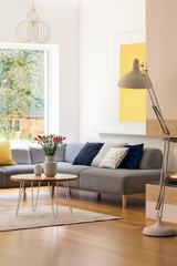 Flowers on table next to grey corner couch in bright living room interior with lamp. Real photo