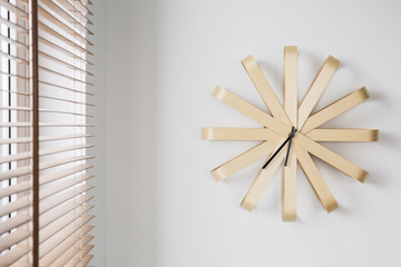 Modern wooden clock on white wall next to window with blinds in simple flat interior. Real photo