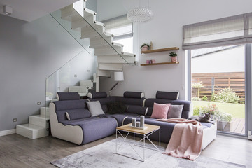 Real photo of an open living room interior with a gray corner sofa next to the window and staircase