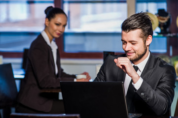 Handsome young man working on laptop and smiling while enjoying coffee in cafe with yang woman in background.