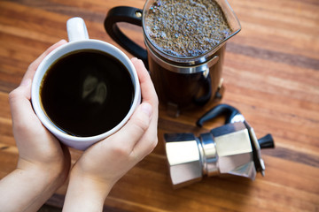 A hand grabbing the handle of a mug of coffee with a french press and smaller pot on a wood cutting...