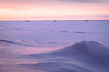 A frozen lake with snow and ice at sunset and snowmobiles in the background.