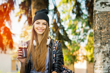Young woman outside on sunny autumn day with headphones, smiling, holding takeaway coffee. Natural lighting, medium retouch.