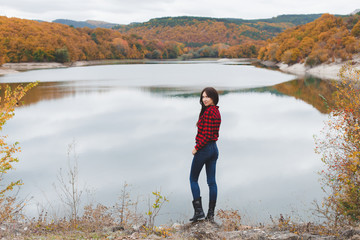 Fototapeta na wymiar Young woman wearing red plaid shirt standing alone near the lake in autumn. Weekend outdoors, cloudy cold weather, fall colors.