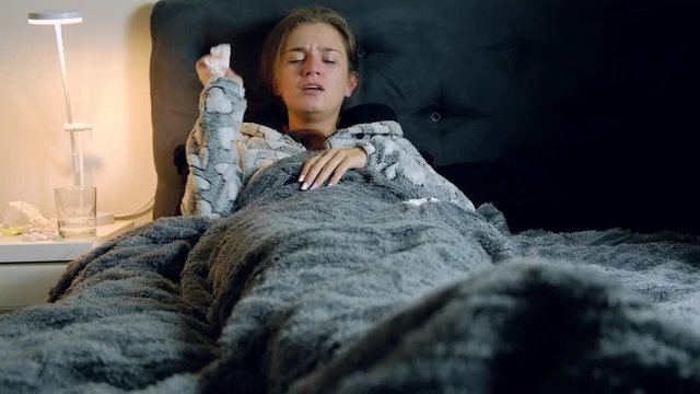 Young sick and tired woman with fever in bed blowing her nose and throwing the tissues. Medicines, thermometer and glass of water on the table.