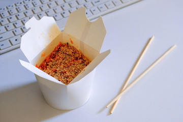 Open cardboard box with Asian food for the programmer. Rice with seafood and sesame seeds and chopsticks on the office table next to a computer keyboard on a white background. Copy space.
