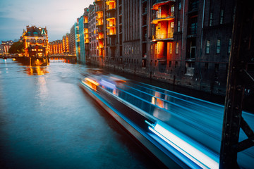 A colorfully illuminated boat cruising on the Wandrahmsfleet at night. The Warehouse District in...