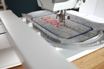 diagonal detail view on a modern computerised sewing machine and embroidery unit with needle down stitching red lettering on grey felt in bright light and a stylish work environment