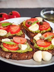 Fototapeta na wymiar Sandwich toasts with tomatoes cherry, mozzarella, avocado, basil and olive oil. Side view on a dark stone dish