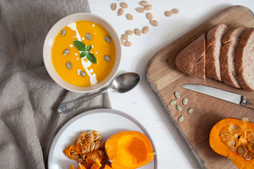 Creamy pumpkin soupwith pumpkin seeds and green, bread with bran and sliced pumpkin on wooden and fabric background. Top view
