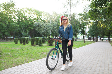 Young happy girl in stylish clothes walking with a green bike in the park and smiles. Evening bike ride along the alley in the park