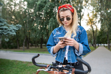 Attractive, serious young girl stands with a bike in the park and uses a smartphone. Stylish girl is biking in the park and using the internet on the smartphone