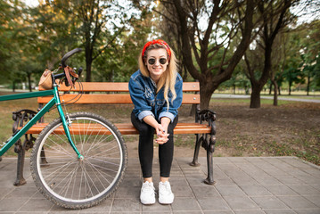 Smiling girl in stylish clothes sitting on a bench in a park with a bike, looking into the camera and smiling. Atractive, positive girl resting on the bench in the park.