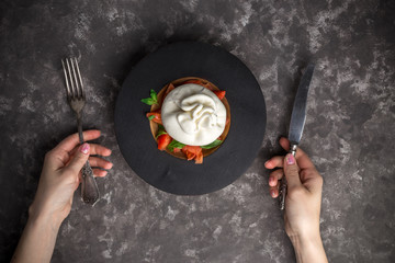Woman eating burrata cheese on small wooden plate served with fresh tomatoes and basil on dark textured background