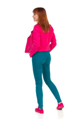 Young Woman In Pink Jacket, Teal Jeans And Sneakers Is Looking Away. Rear View.