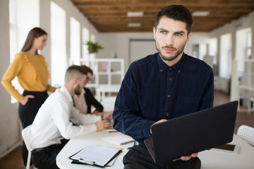 Young serious man with beard in shirt thoughtfully looking aside holding laptop in hands while spending time in office with colleagues on background