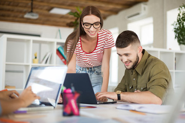Young cheerful man in shirt and woman in striped T-shirt and eyeglasses working together with laptop. Creative business people spending time at work in modern office