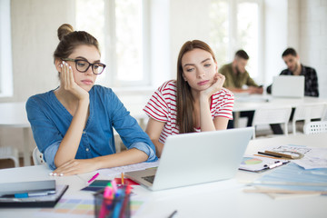 Serious girls leaning on hands while thoughtfully working with laptop. Young women spending time in modern office together