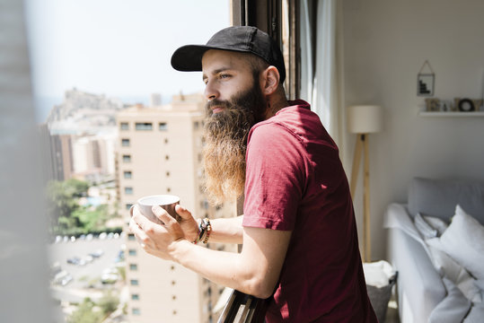 Thoughtful bearded man looking through window while holding coffee cup at home